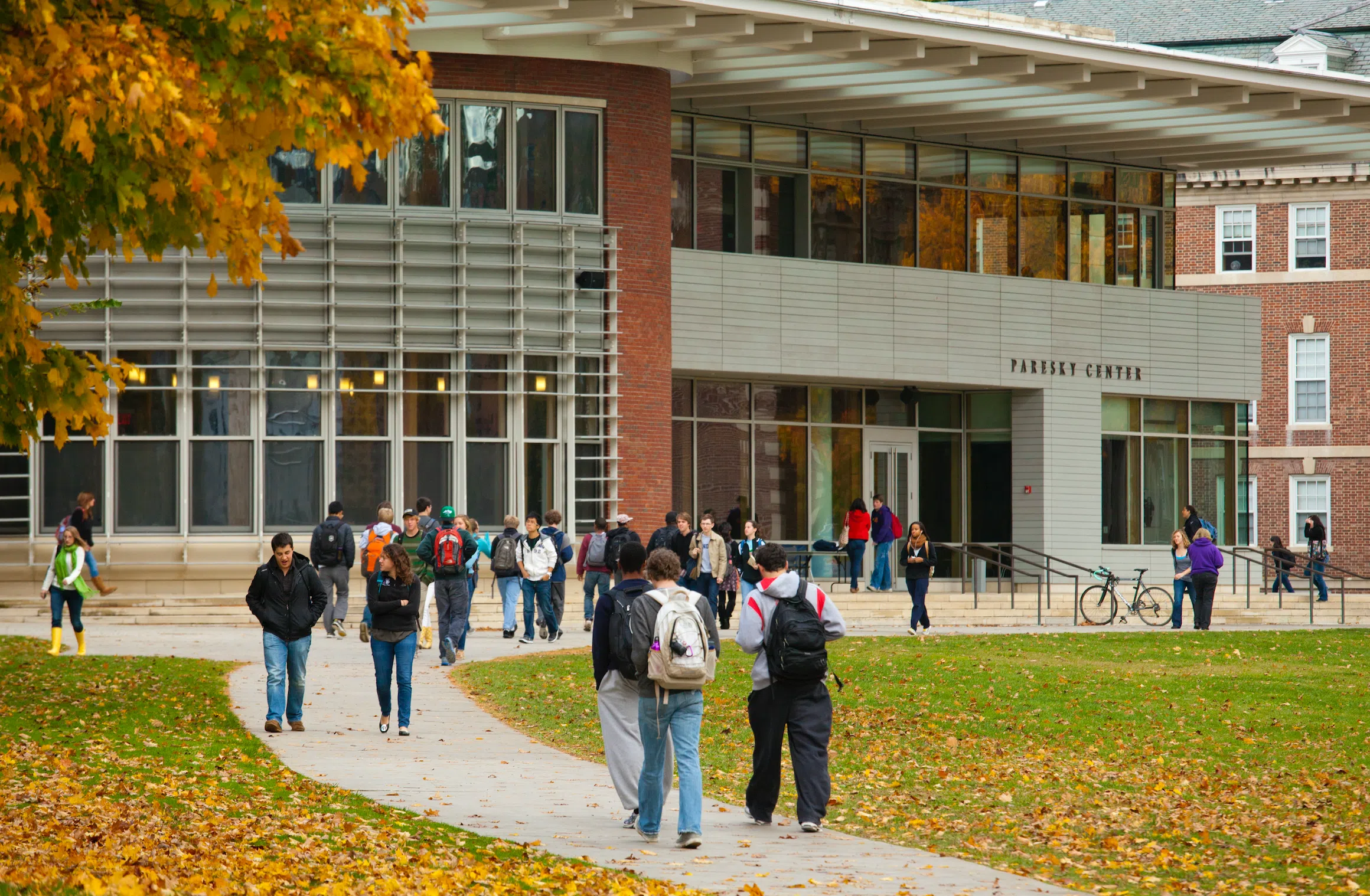 An image of students walking on a sidewalk toward Pareksy Center, a large modern building with large glass windows, steps, and silver and red brick finishes.